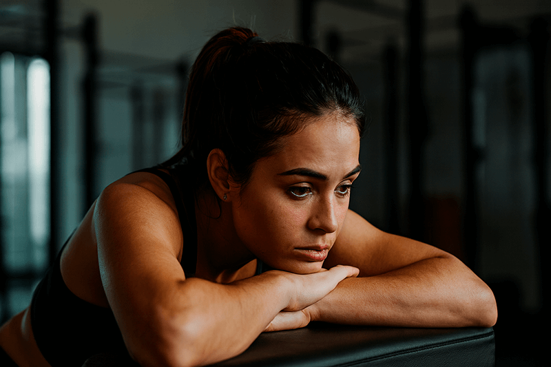 Уставшая молодая тренер в спортивном зале сидит, задумчиво опершись на руки, — визуал отражает эмоциональное выгорание и усталость новичков в фитнес-профессии.
