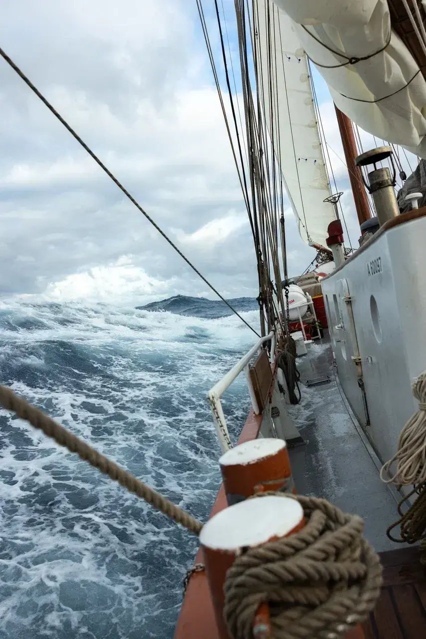 The deck of a traditional expedition schooner battling the powerful waves of the legendary Drake Passage. Towering swells crash alongside the vessel under dramatic storm clouds, capturing the raw power of one of the world's most challenging and iconic ocean crossings between South America and Antarctica.