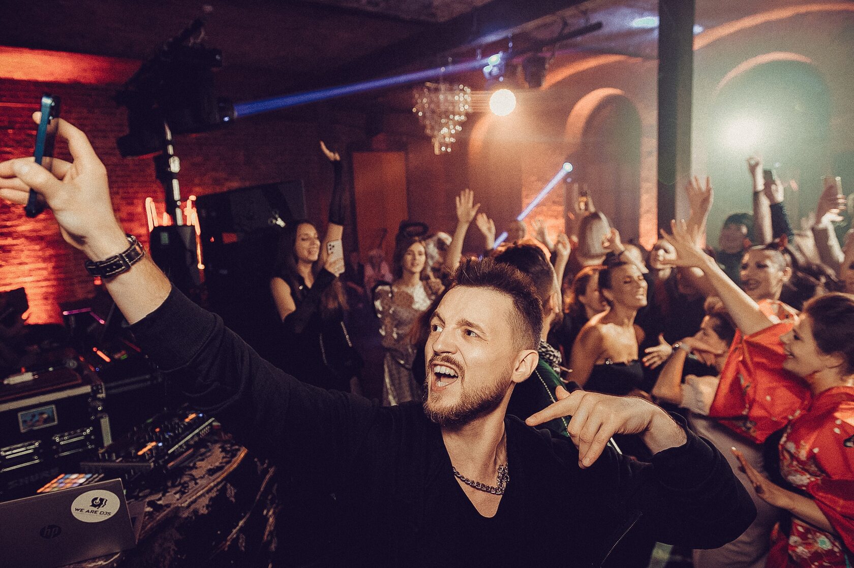 DJ ROZHKOV на переднем плане улыбается и поднимает руку на фоне танцующей компании гостей.