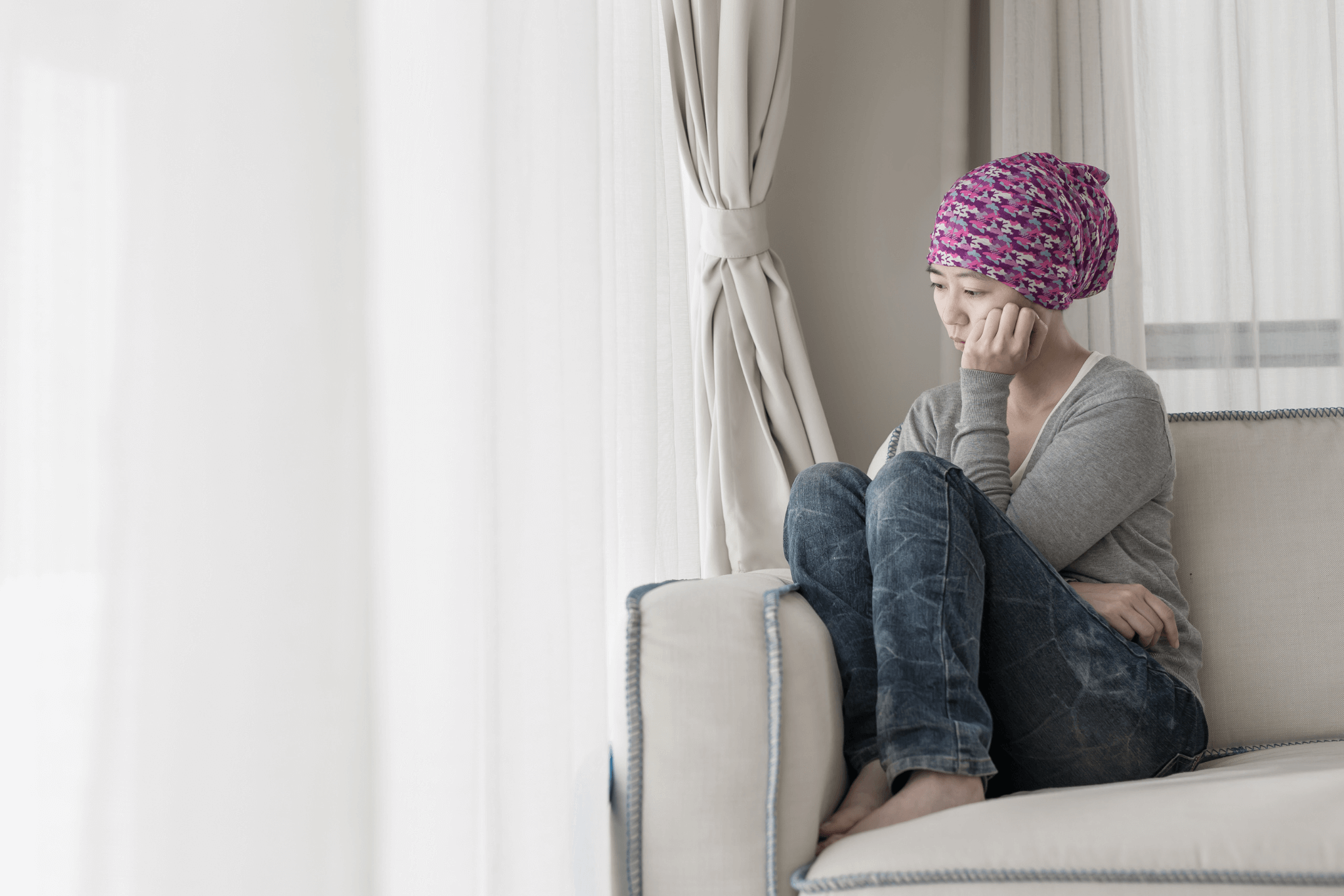 Traurig blickende Tumorpatientin sitzt mit Kopftuch in der Ecke eines Sofas