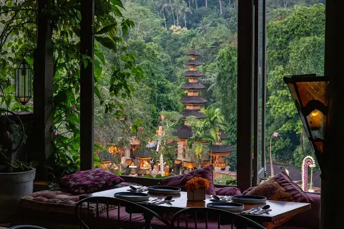 A picturesque spot for a top dinner in Ubud, showing a cozy dining table with plush cushions looking directly out over a beautifully illuminated traditional Balinese temple.