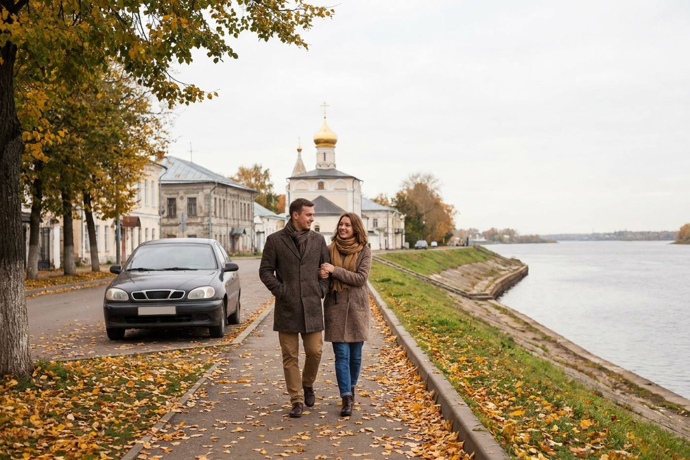 Мужчина и женщина в пальто идут под руку по усыпанной желтыми листьями набережной на фоне старинного храма и реки