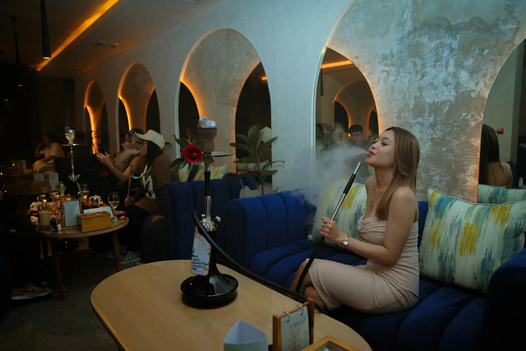 An elegant woman enjoying a smooth shisha smoke session on a plush blue couch inside the beautifully arched interior of Eden Hookah Club Ubud.
