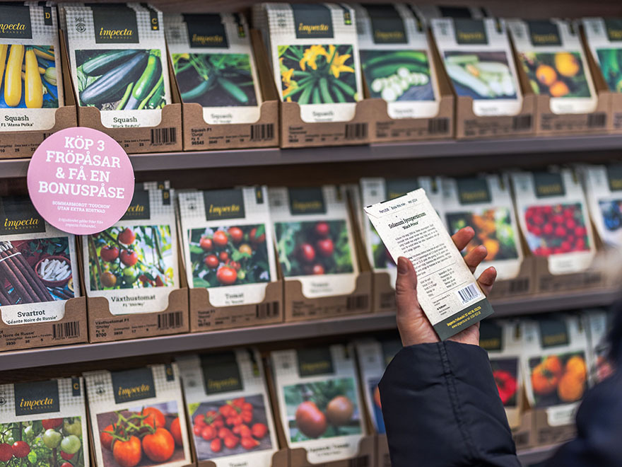 Hand holding seed packet in front of organized display of various vegetable seed packets, including tomatoes, squash, and beans, in a garden center setting, promoting gardening supplies for home gardeners.
