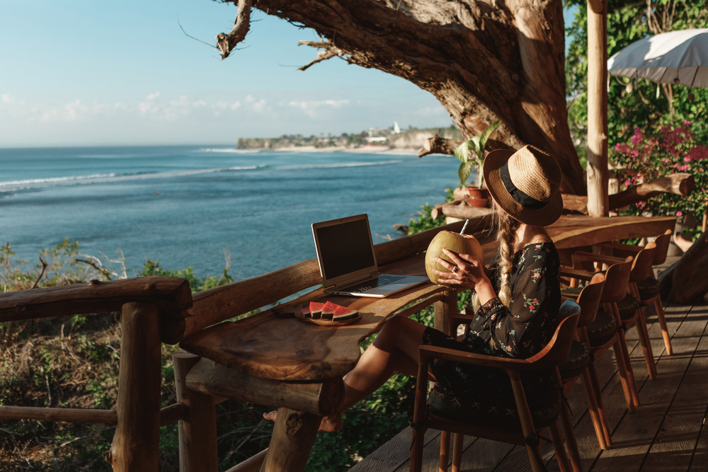 A serene photograph of a woman wearing a straw hat working on her laptop at a wooden cliffside bar overlooking the ocean in Uluwatu. This visual captures the dream lifestyle many seek when starting as a digital nomad in Bali.