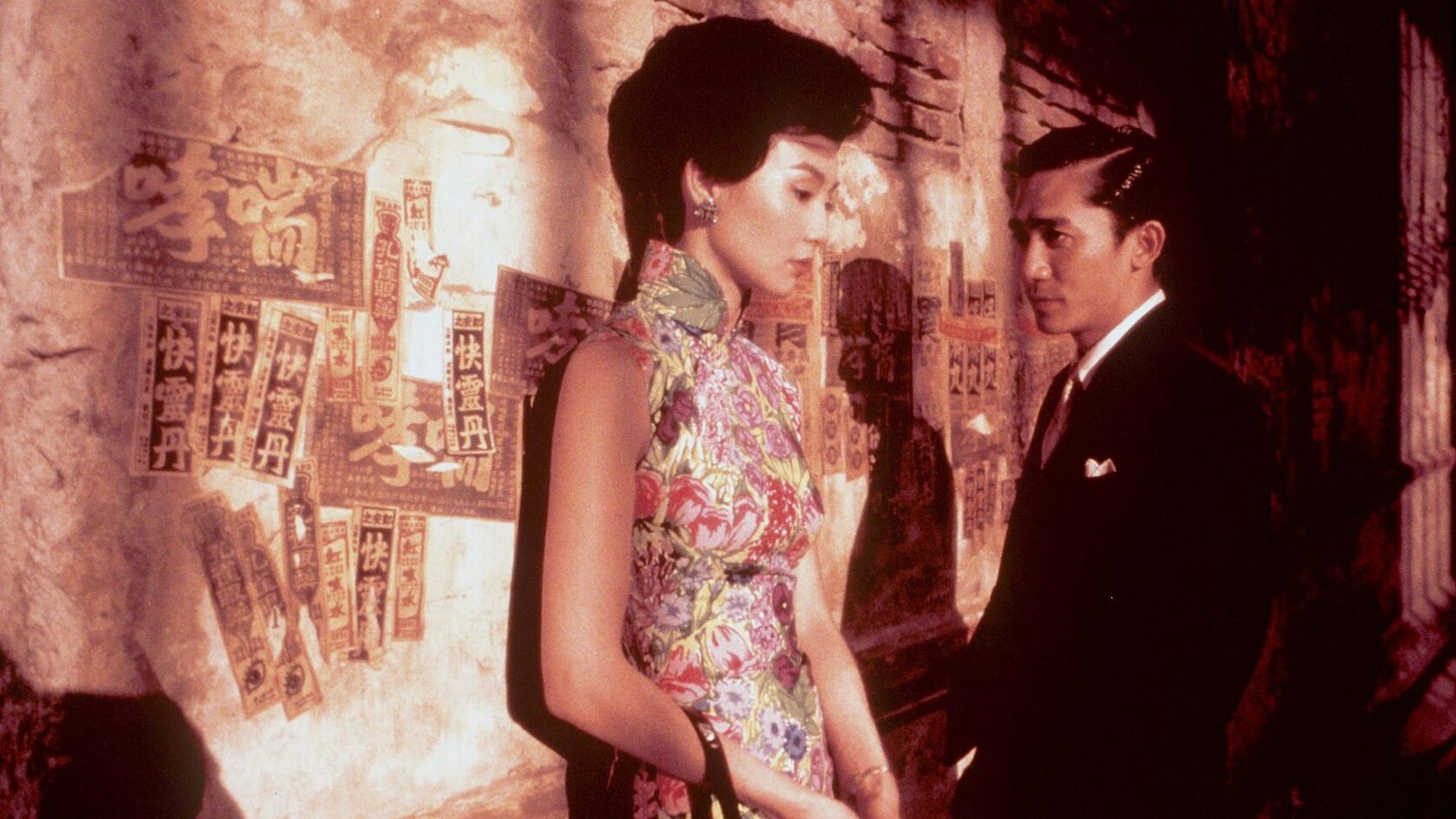 In the Mood for Love: Chou Mo-wan looks at Su Li-zhen as she leans against a wall covered in faded flyers. These two need to kiss already.