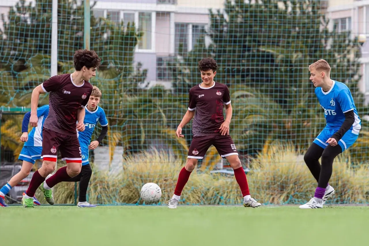 Игроки в бордовой и синей форме рядом с мячом на футбольном поле