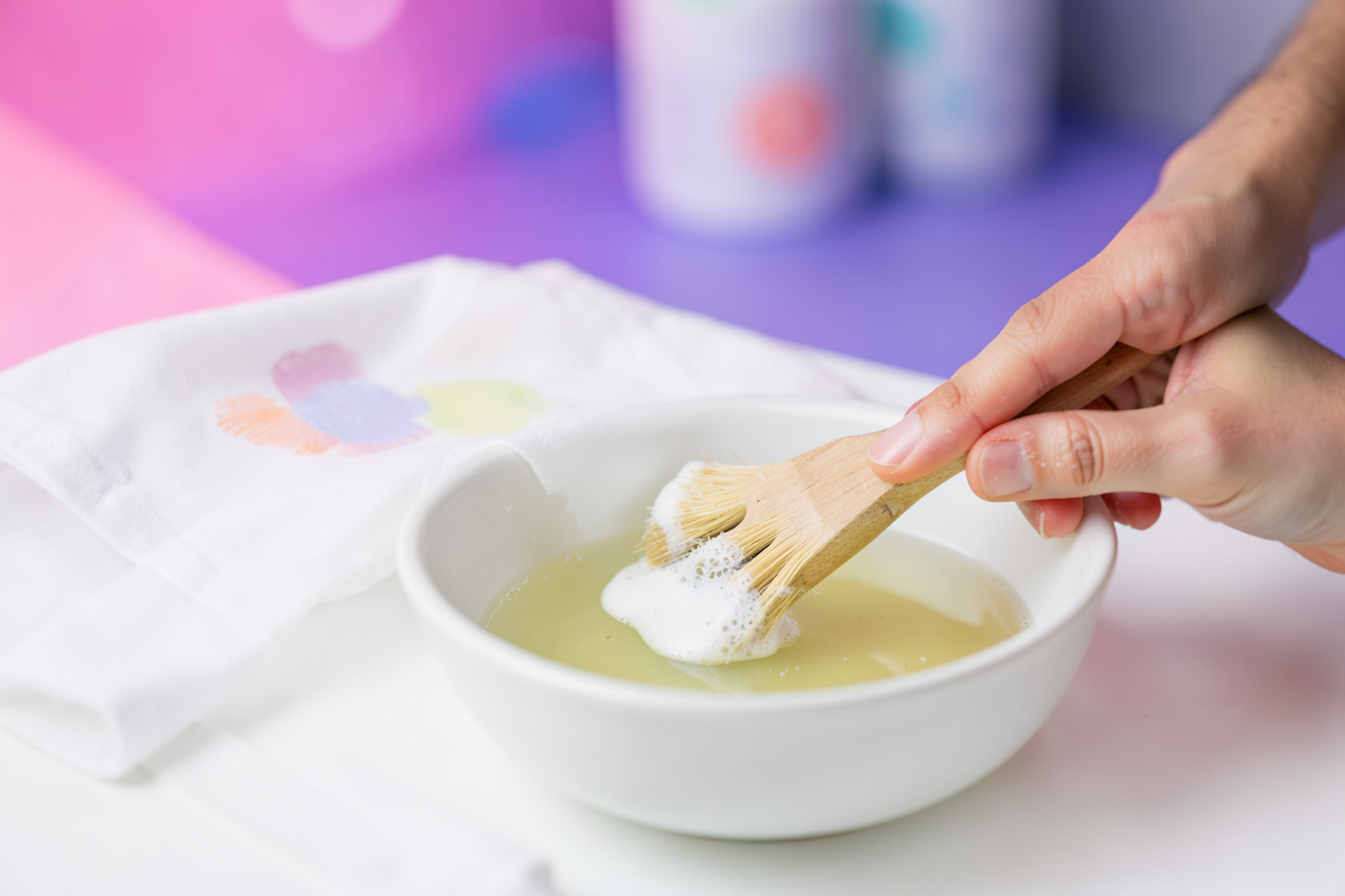 Close-up of a hand treating a stain on a white shirt with gentle soap and a soft brush over a bowl of warm water, stain visibly being lifted, no product labels