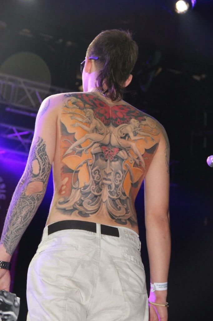 A massive full-back color tattoo featuring a symmetrical neo-traditional design with a central face, mythical figures, and a red butterfly under stage lighting