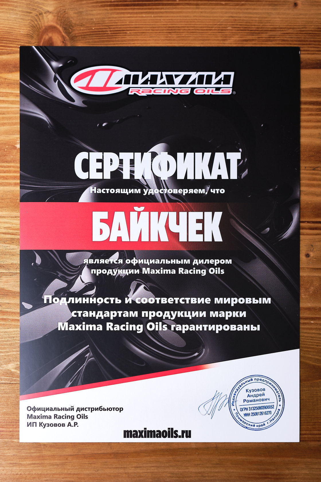 Сертификат официального дилера Maxima Racing Oils для мастерской Байкчек на деревянном столе