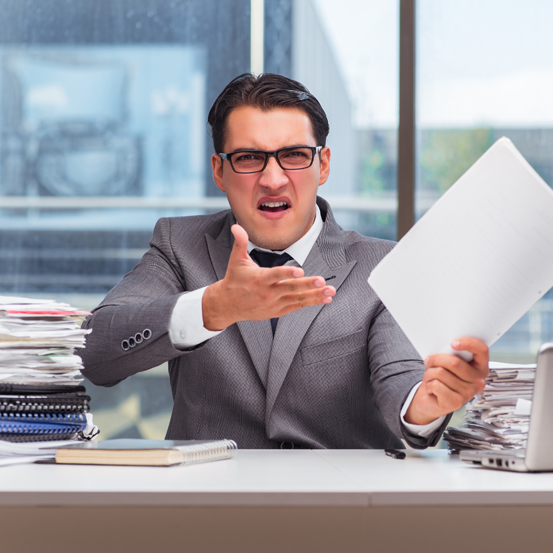 Caucasian man at desk with hand up holding a paper with angry expression