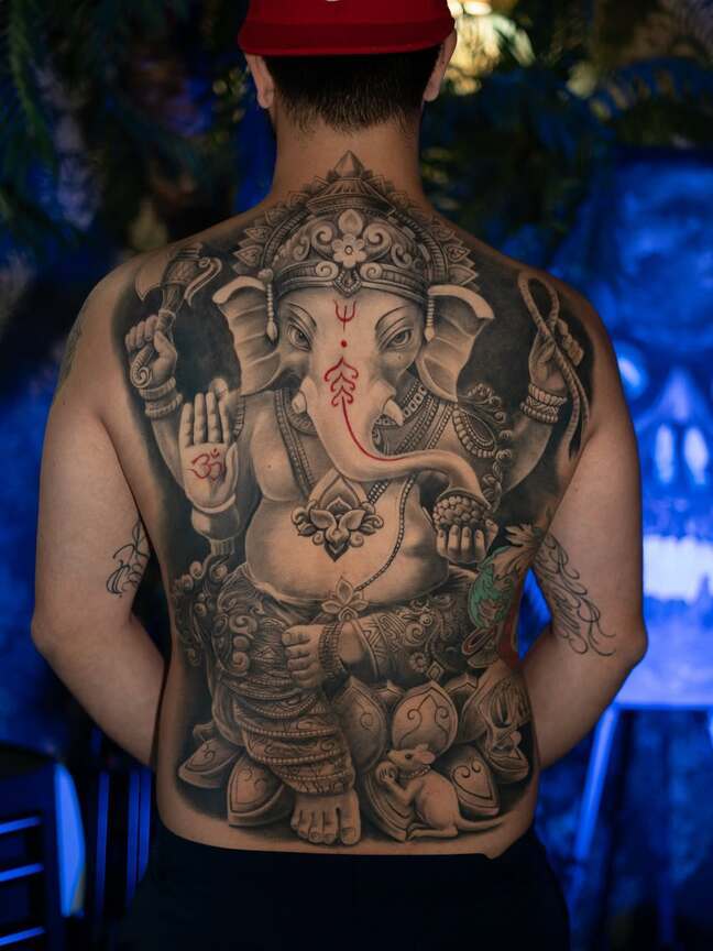 A healed, large-scale black and grey realism tattoo of the Hindu deity Ganesha covering the entire back, featuring intricate ornamental details, sacred symbols, and a red Om sign on the palm, by Rinat Mingazdinov, Los Angeles