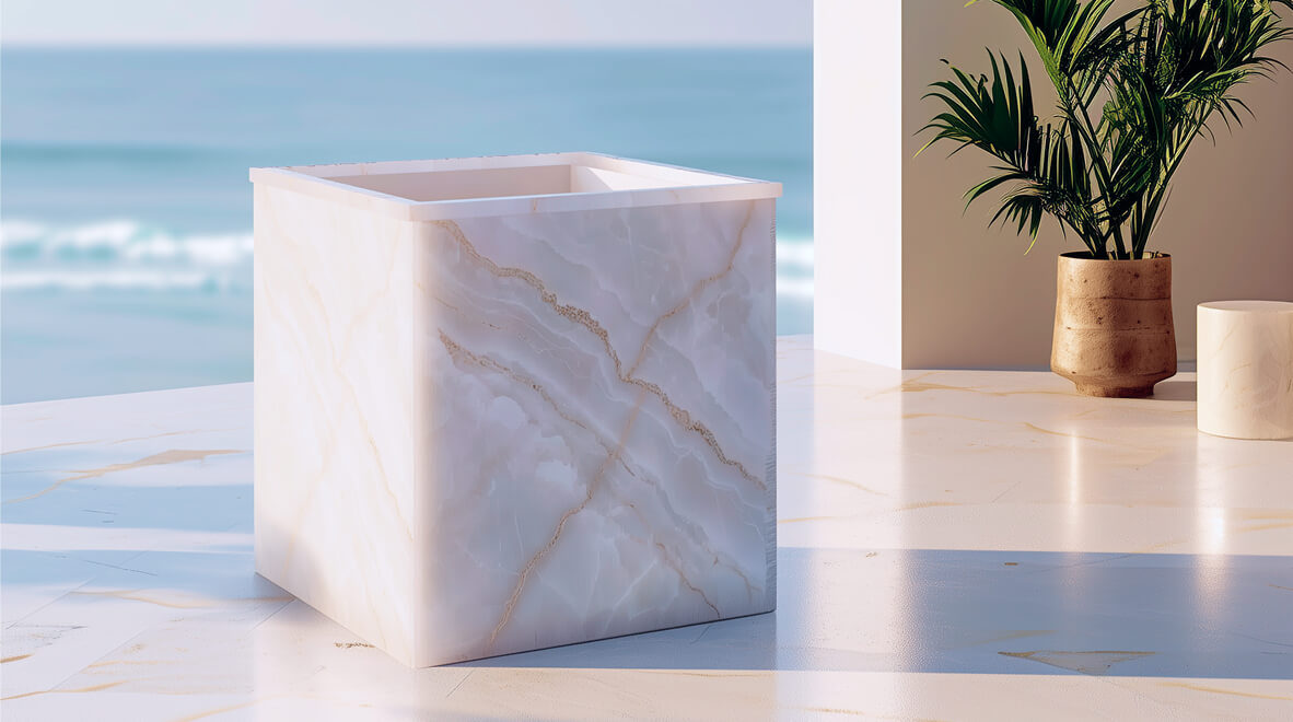 Luxury cube-shaped onyx cold plunge or ice bath tub by Arctic Bear Wellness, featuring premium translucent white marble stone panels with golden-brown veining, installed on a minimalist marble patio overlooking a calm turquoise ocean background with soft beach waves.