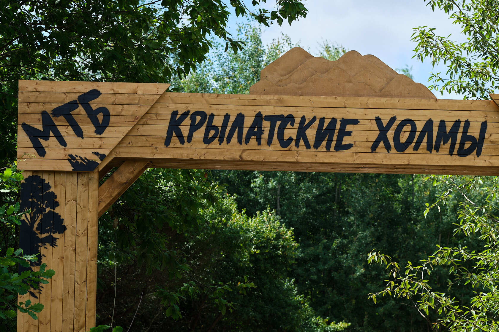 Деревянная входная арка с надписью «МТБ Крылатские холмы» на фоне зеленых деревьев