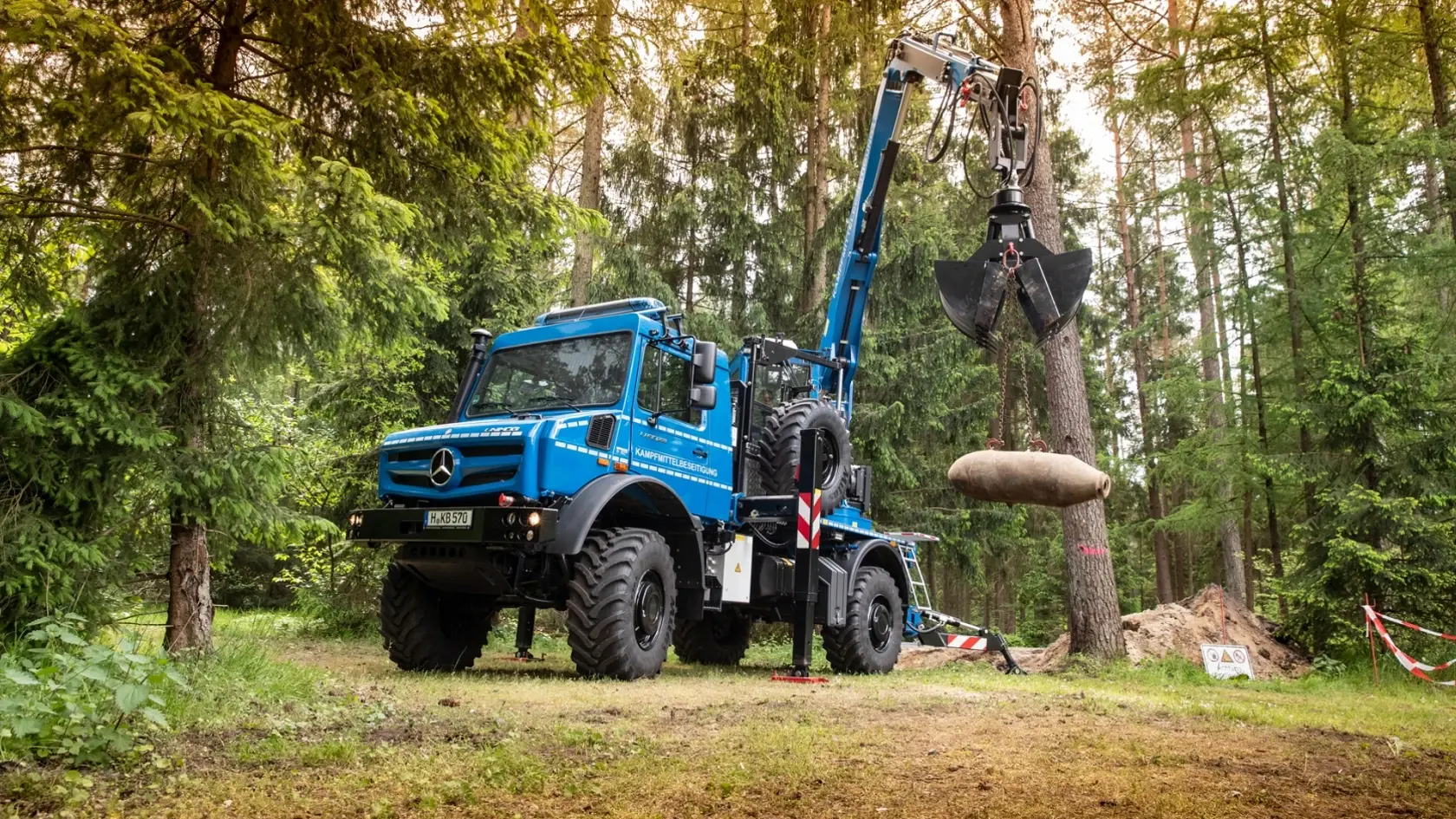 Синий Unimog с краном-манипулятором поднимает крупный цилиндрический груз в лесу