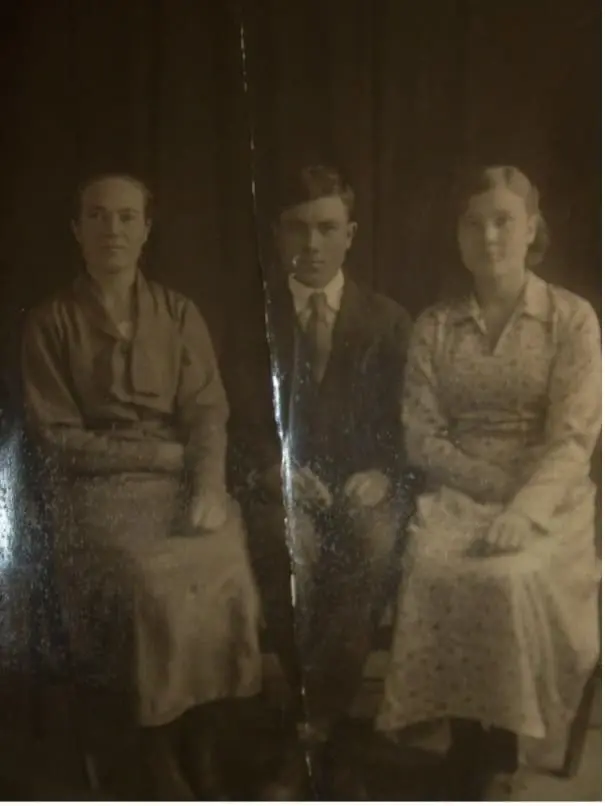 Архивная чёрно-белая фотография первой половины XX века: молодой мужчина в тёмном костюме и галстуке сидит между двумя молодыми женщинами в длинных платьях. Снимок сделан в фотостудии на тёмном фоне; на фотографии заметны вертикальный разрыв, потёртости и следы времени.