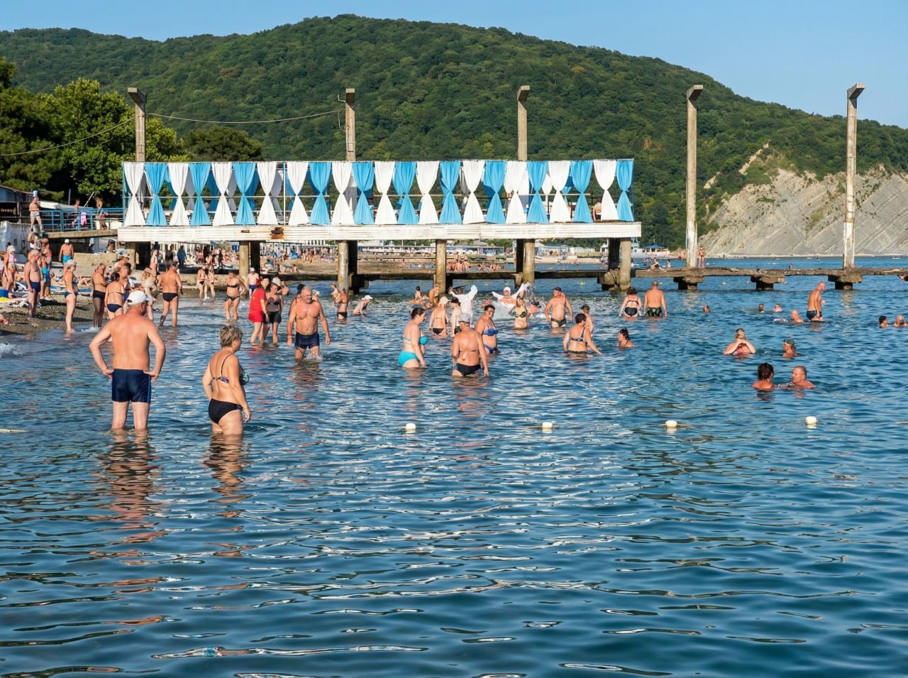 Детский пляж у моря с мелкой водой, купающимися людьми и пирсом на фоне гор
