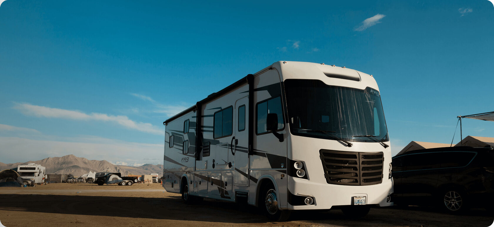 A large RV parked at the Burning Man festival at dusk, with neon art installations and the desert landscape.
