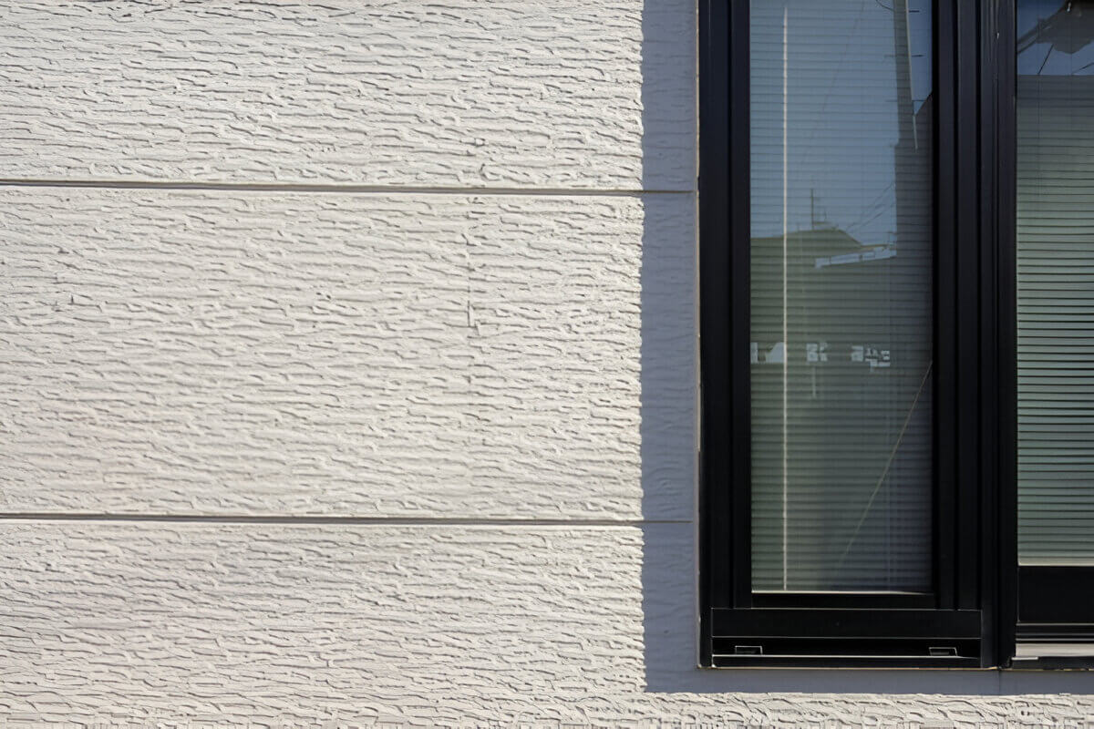 Фрагмент фасада с декоративной штукатуркой светлого оттенка. Видна фактура штукатурного покрытия и примыкание к оконному проёму с тёмной рамой.