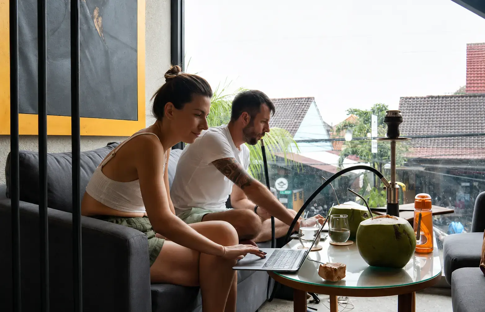 A relaxed daytime scene of two people casually working on their laptops while sharing a couch. Fresh coconuts are on the table, and a hookah stands nearby. A large window looks out over rooftops, showcasing the hybrid workspace vibe at Eden Hookah Club, popular with the digital nomad in Bali community.