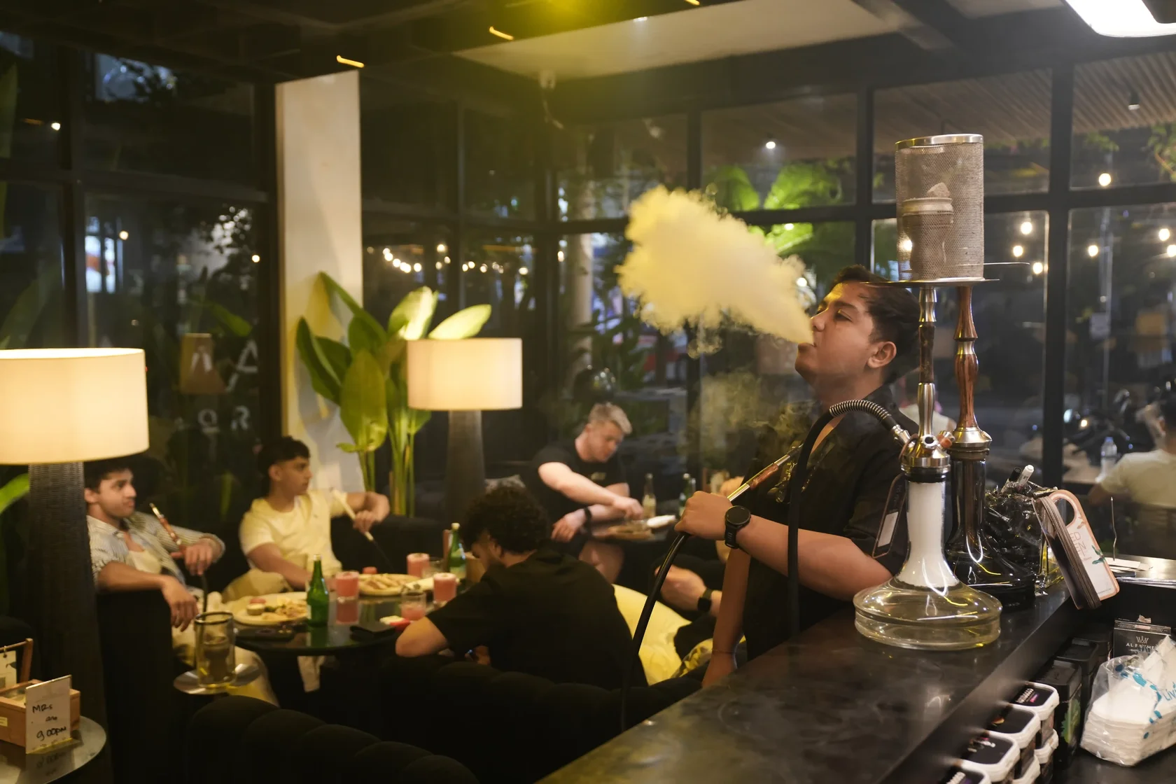 A vibrant shot of guests socializing and enjoying hookahs in a modern plant-filled indoor lounge with warm lighting in Bali capturing the lively midweek nightlife scene.
