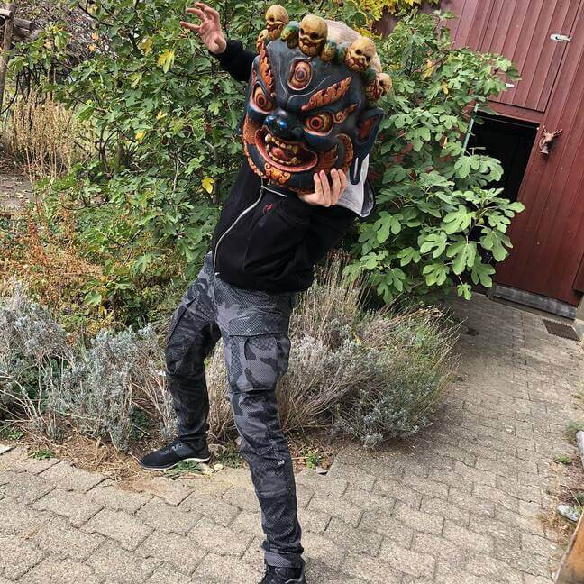 Master Rinat, wearing a black sweatshirt and camouflage pants, holds a Tibetan mask of the protector Mahakala with skulls to his face against a garden backdrop.