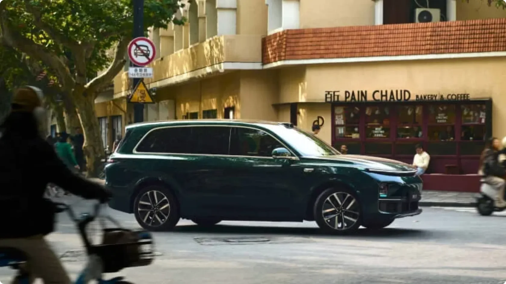 Гибридный кроссовер Li Auto в глубоком зеленом цвете с лидаром на крыше, движущийся по улице города на фоне кафе и велосипедиста.