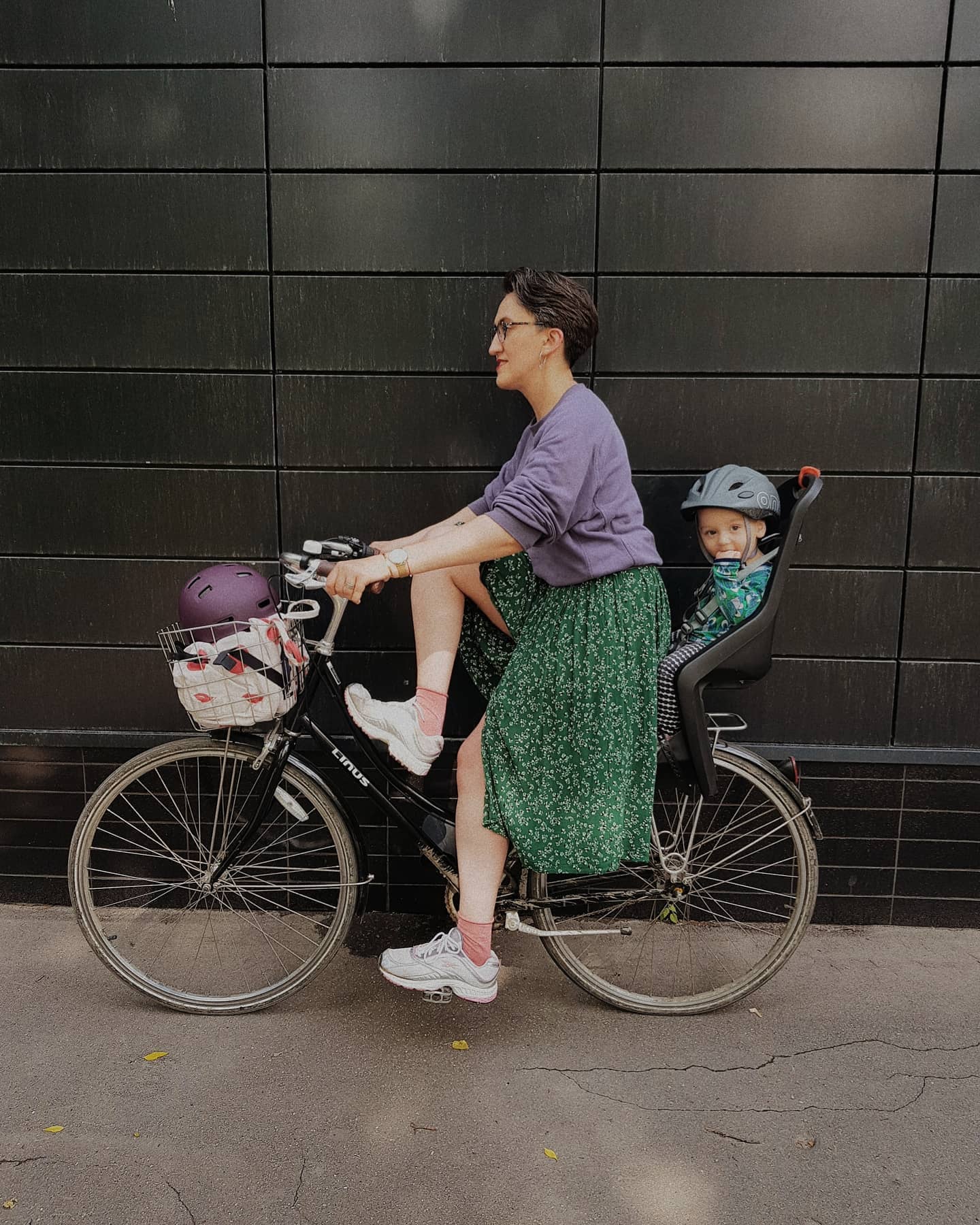 девушка в зеленой юбке едет на велосипеде с ребенком на багажнике 