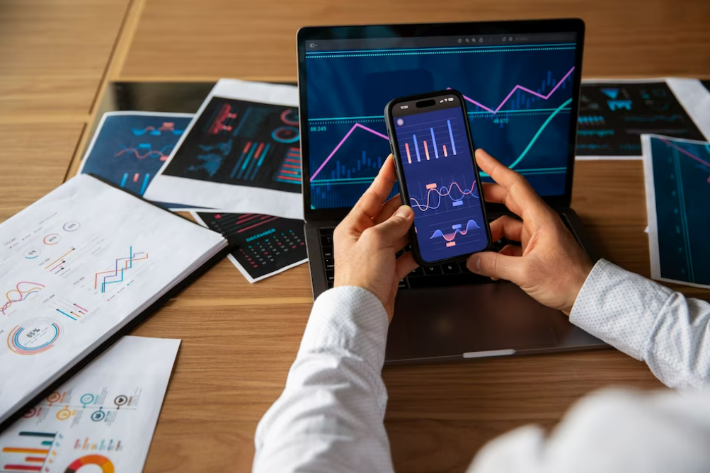 A person holding a mobile phone, in front of a laptop with crypto chart patterns. On a table with printed charts on sheets of paper