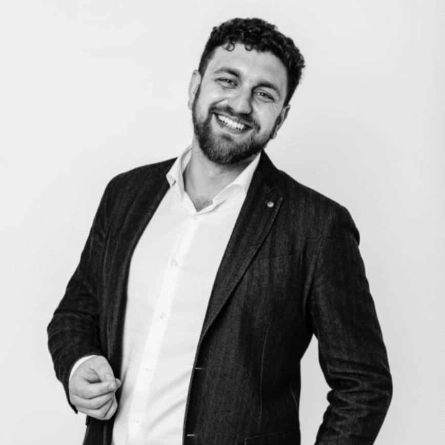Монохромная портретная фотография улыбающегося мужчины с темными вьющимися волосами и небольшой бородкой, в темном пиджаке и белой рубашке, смотрящего прямо в камеру, на фоне светлой однотонной стены.
