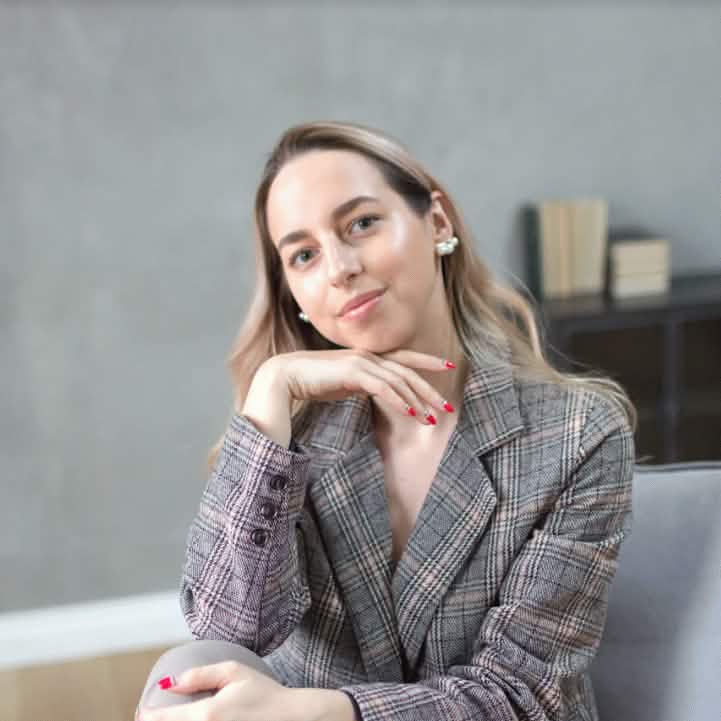 Портретная фотография молодой женщины со светлыми вьющимися волосами, с легким макияжем и красными ногтями, в сером клетчатом пиджаке, сидящей на стуле, подпирающей подбородок рукой, на фоне размытой серой стены с книгами на полке.