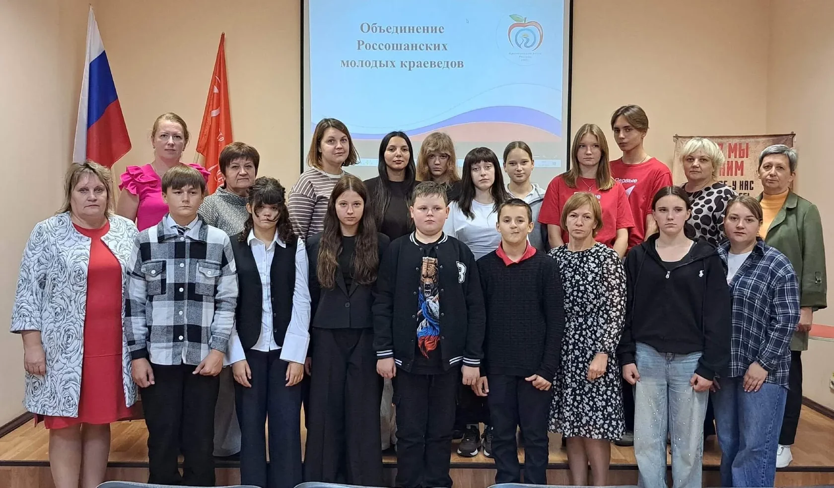 На фото — групповое фото участников мероприятия «Объединение Россошанских молодых краеведов». В кадре — школьники, педагоги и кураторы, выстроенные в два ряда. На фоне — проекционный экран с названием объединения и его логотипом, слева видны флаги (в том