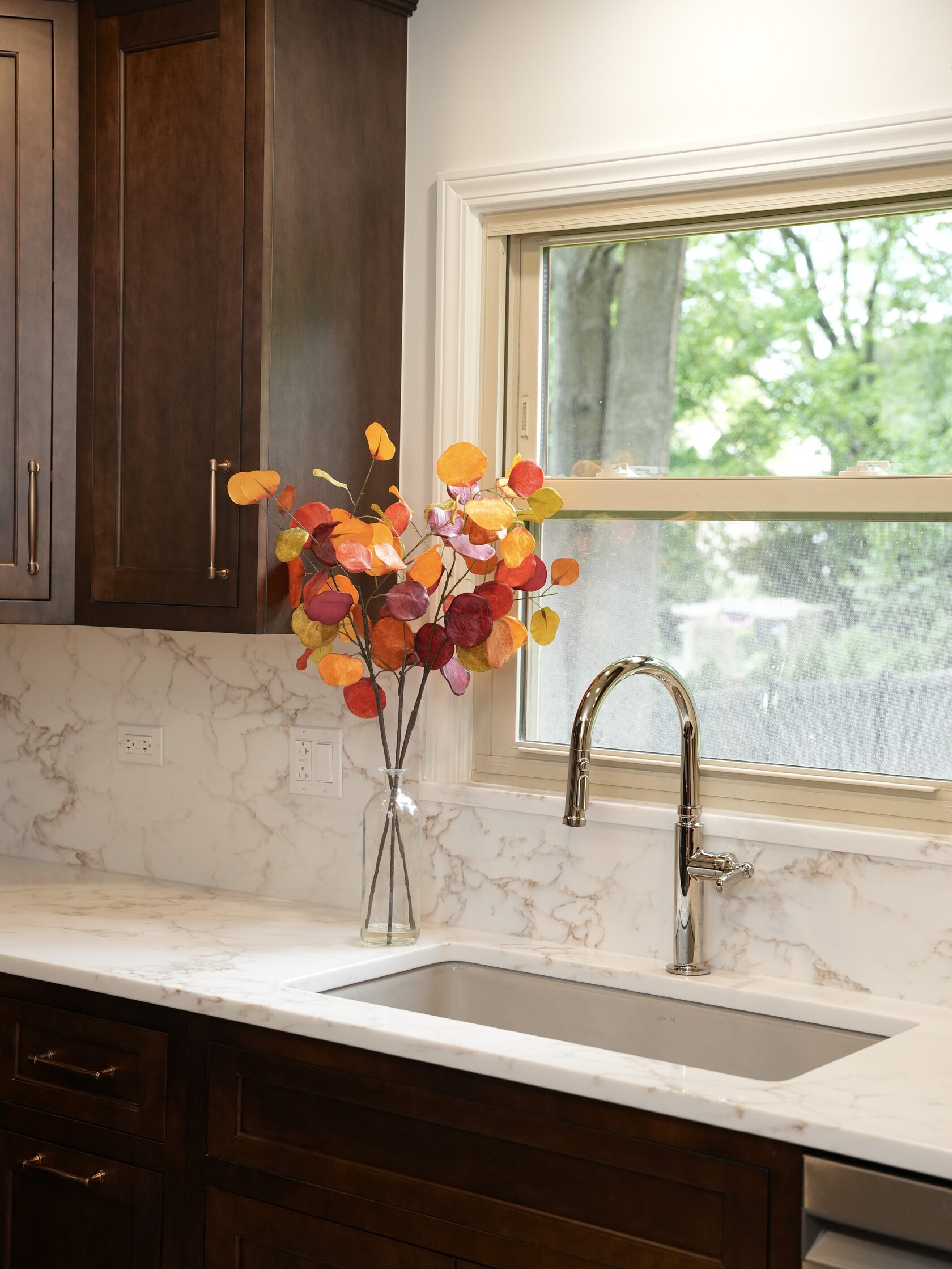 Название: The window-centered sink area brings natural light into the work zone and softens the darker cabinetry with a bright, open focal point. - описание: Close-up of a kitchen sink under a window, with dark cabinetry, a chrome faucet, light stone-look counter, and colorful stems in a glass vase.