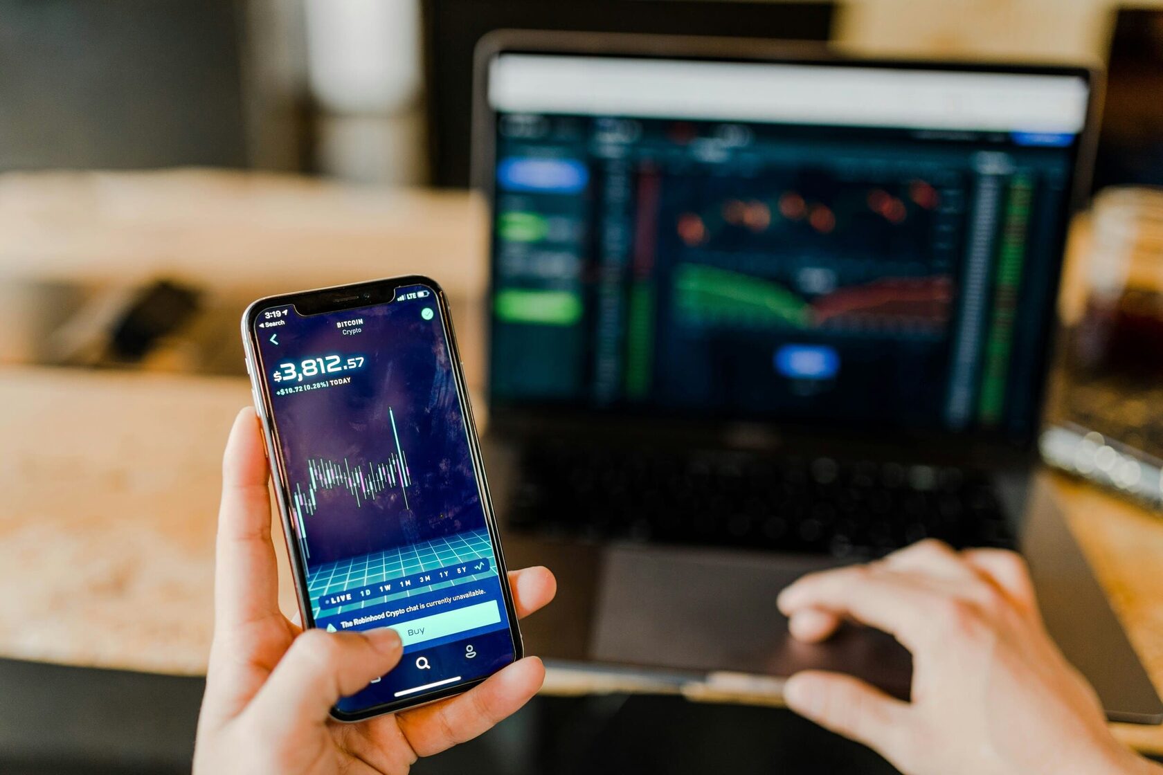 Retail investor monitoring portfolio balance on a mobile trading app with a laptop dashboard in the background.