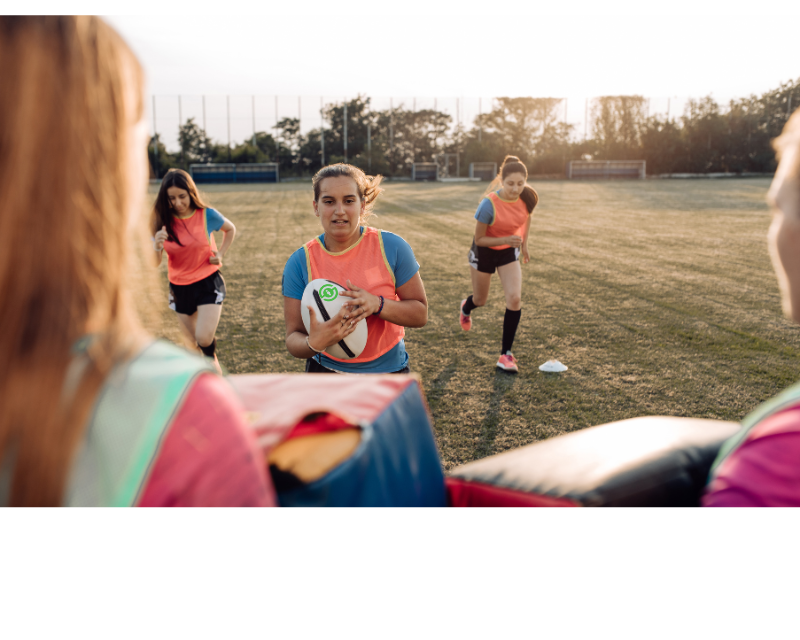 Girls training for rugby, one is carrying the ball and running towards a tackle bag being held by another girl