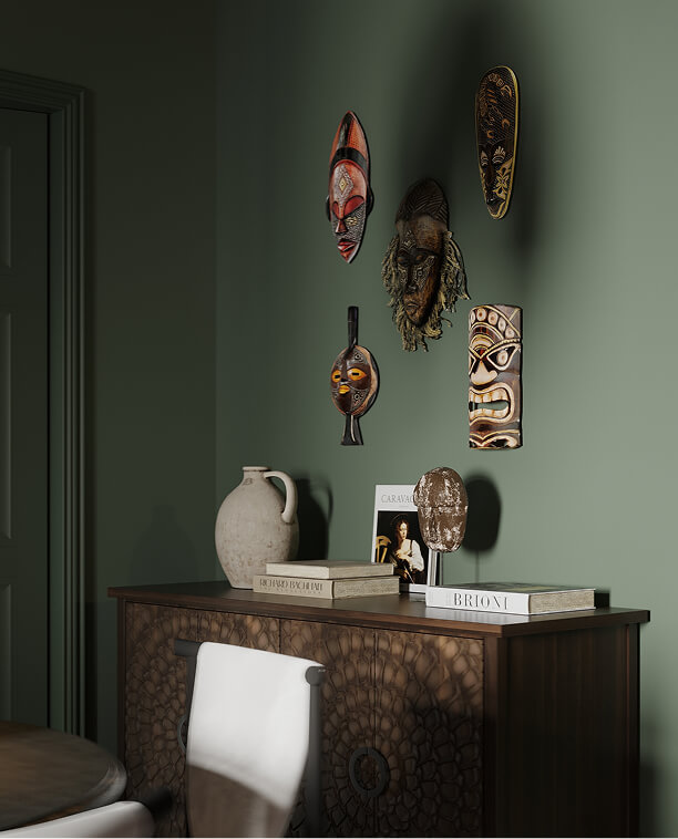 Деревянный комод с резными узорами, на котором стоят книги, керамический кувшин и декоративные предметы. Над комодом на зеленой стене висят несколько этнических масок с яркими узорами и различными текстурами