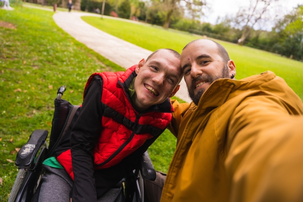 Two men in a park taking a selfie. One is in a wheelchair and red puffer gilet, the other in an orange puffer jacket