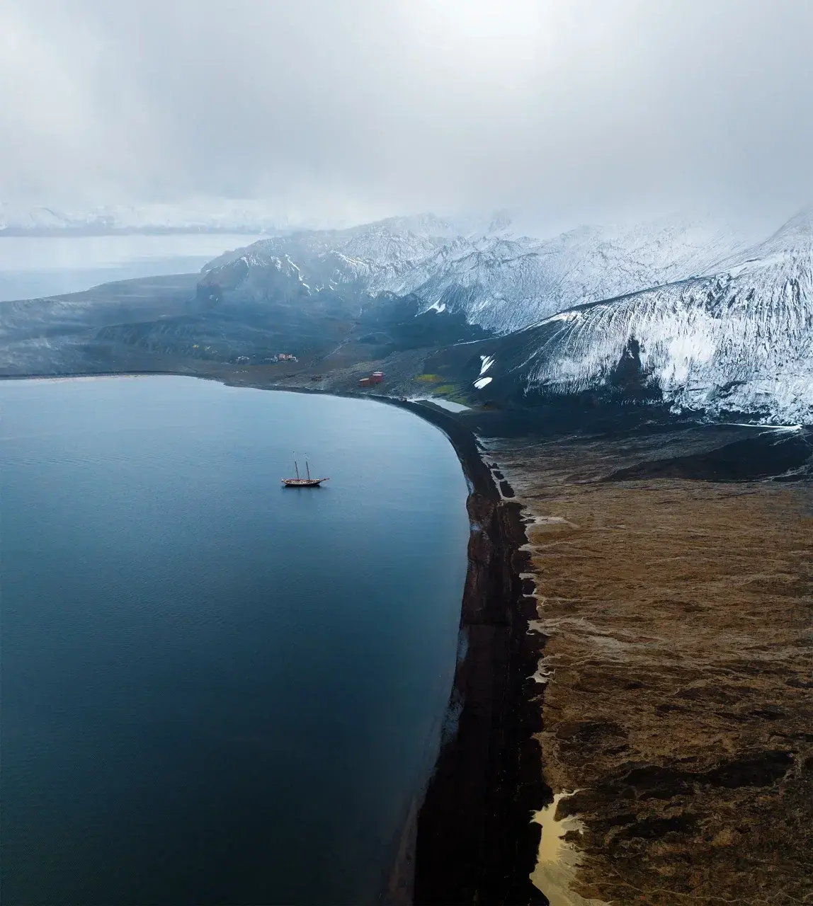 View from an airplane window over a remote Arctic coastline with a black volcanic beach calm bay snow-covered mountains and a solitary sailing yacht anchored near the shore beneath low clouds.