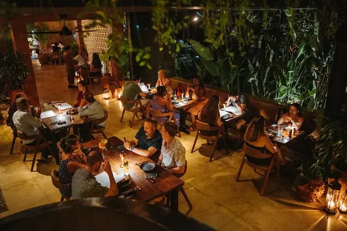 Diners having a fantastic top dinner in Ubud on a beautifully lit outdoor patio, surrounded by tropical greenery and a sophisticated, warm community vibe.