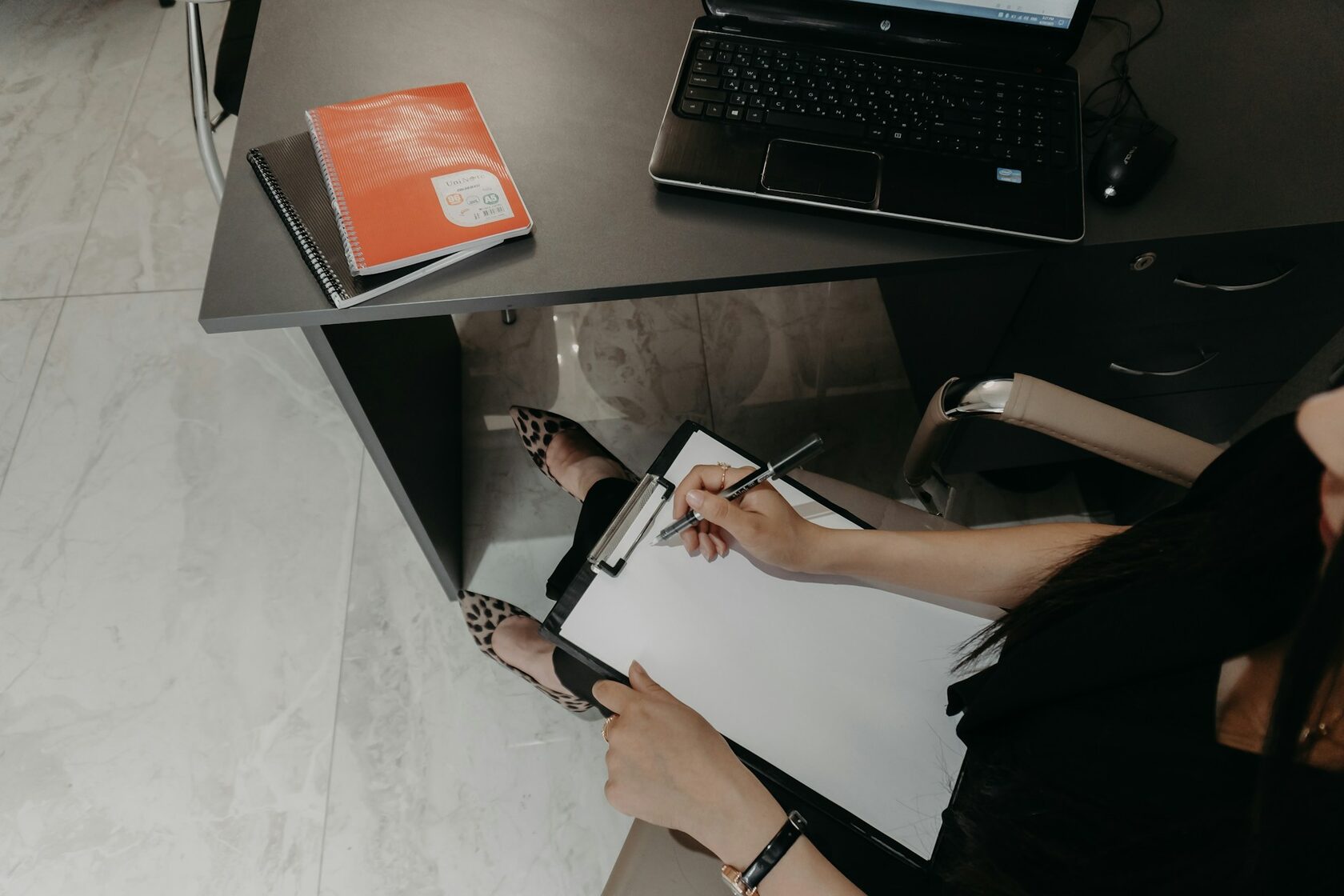 Person writing on clipboard at desk with laptop