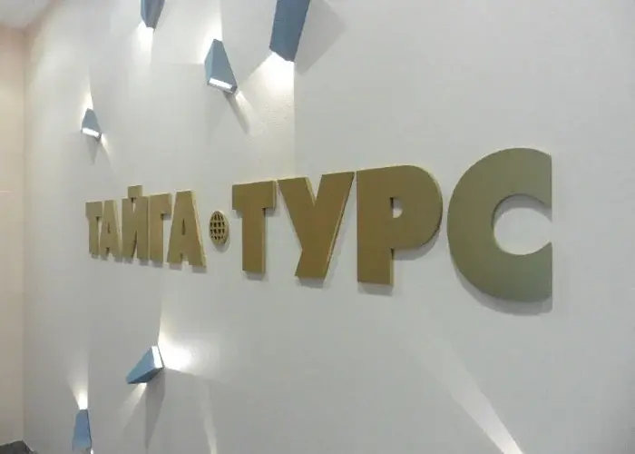 Золотистая вывеска НАТА ТУРС на белой стене с направленным освещением