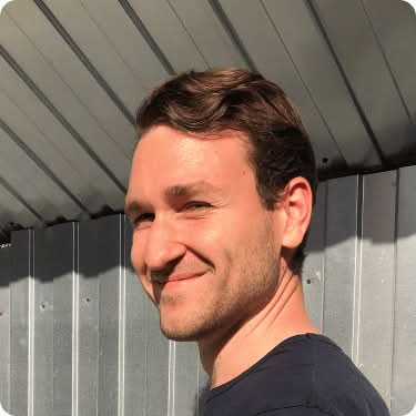 Портретная фотография молодого мужчины с волнистыми светло-каштановыми волосами и легкой небритостью, с легкой улыбкой и смотрящего вправо, на фоне металлической гофрированной поверхности.