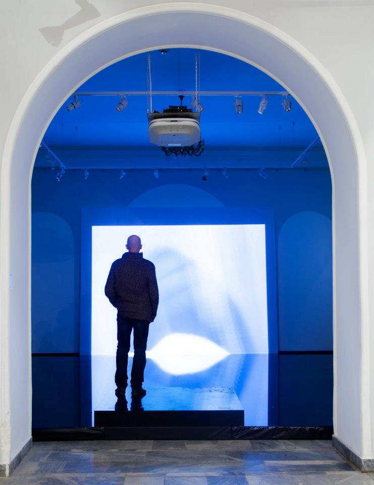 A solitary figure standing inside a luminous blue arched room with a video projection on the floor