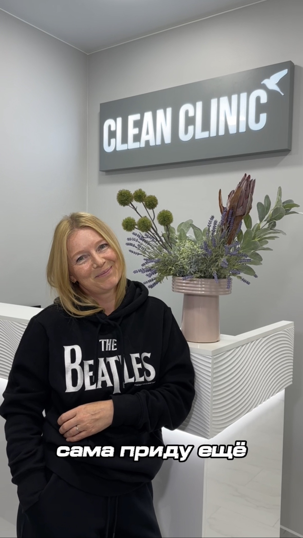 Молодая женщина в черном свитшоте с надписью Beatles на фоне цветов и логотипа Clean Clinic рассказывает отзыв о приеме врача и капельнице