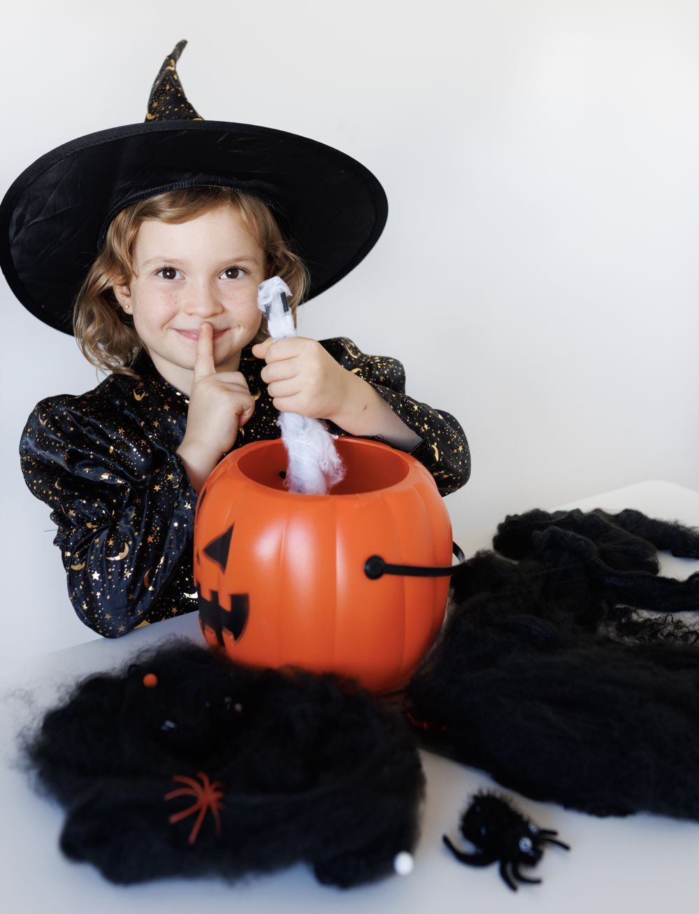 Una niña sonriente con sombrero de bruja, con el dedo en los labios, junto a un cubo-calabaza