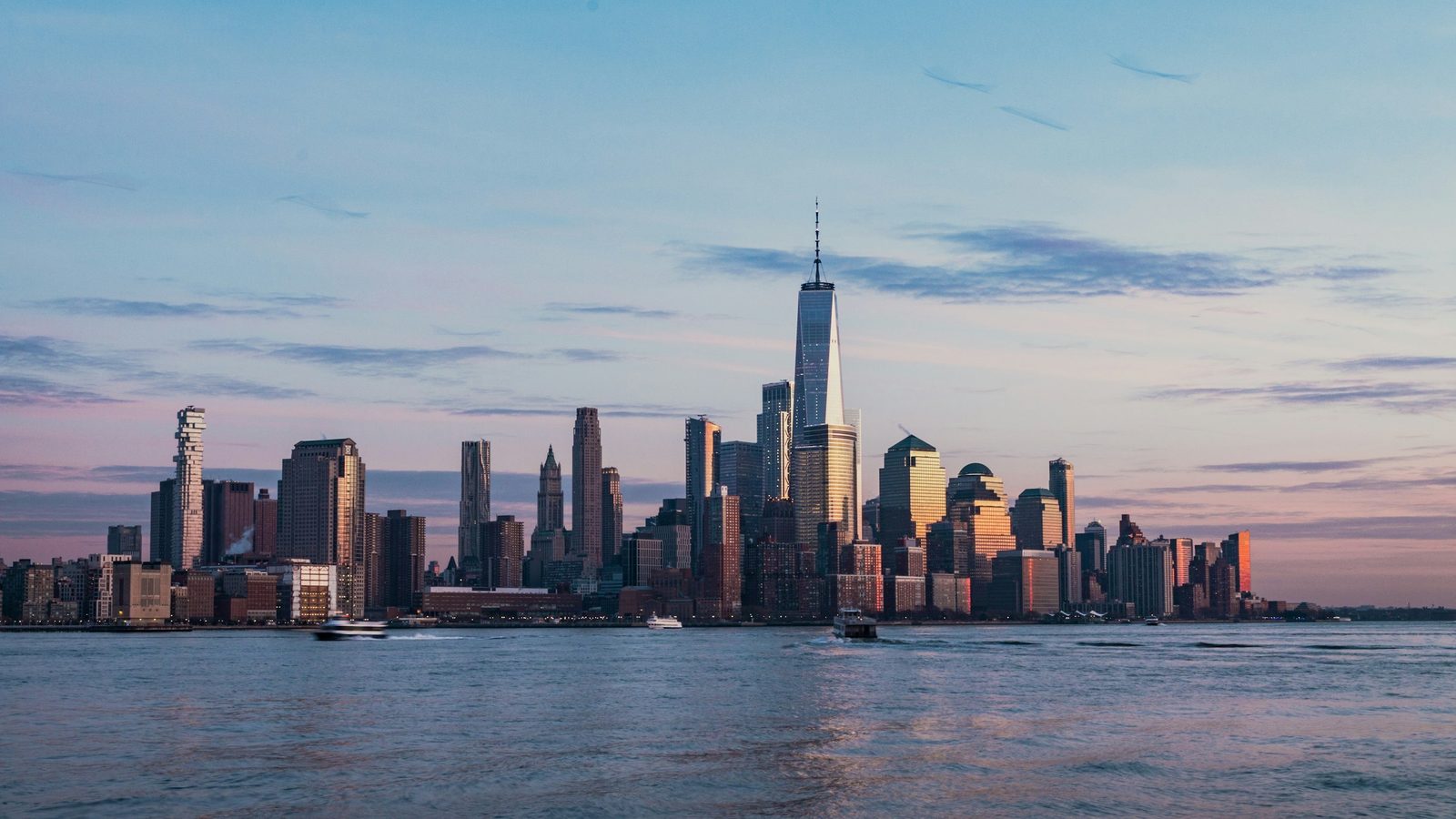 The Lower Manhattan skyline at dusk seen across the Hudson River, with One World Trade Center in the middle and ferries on the water