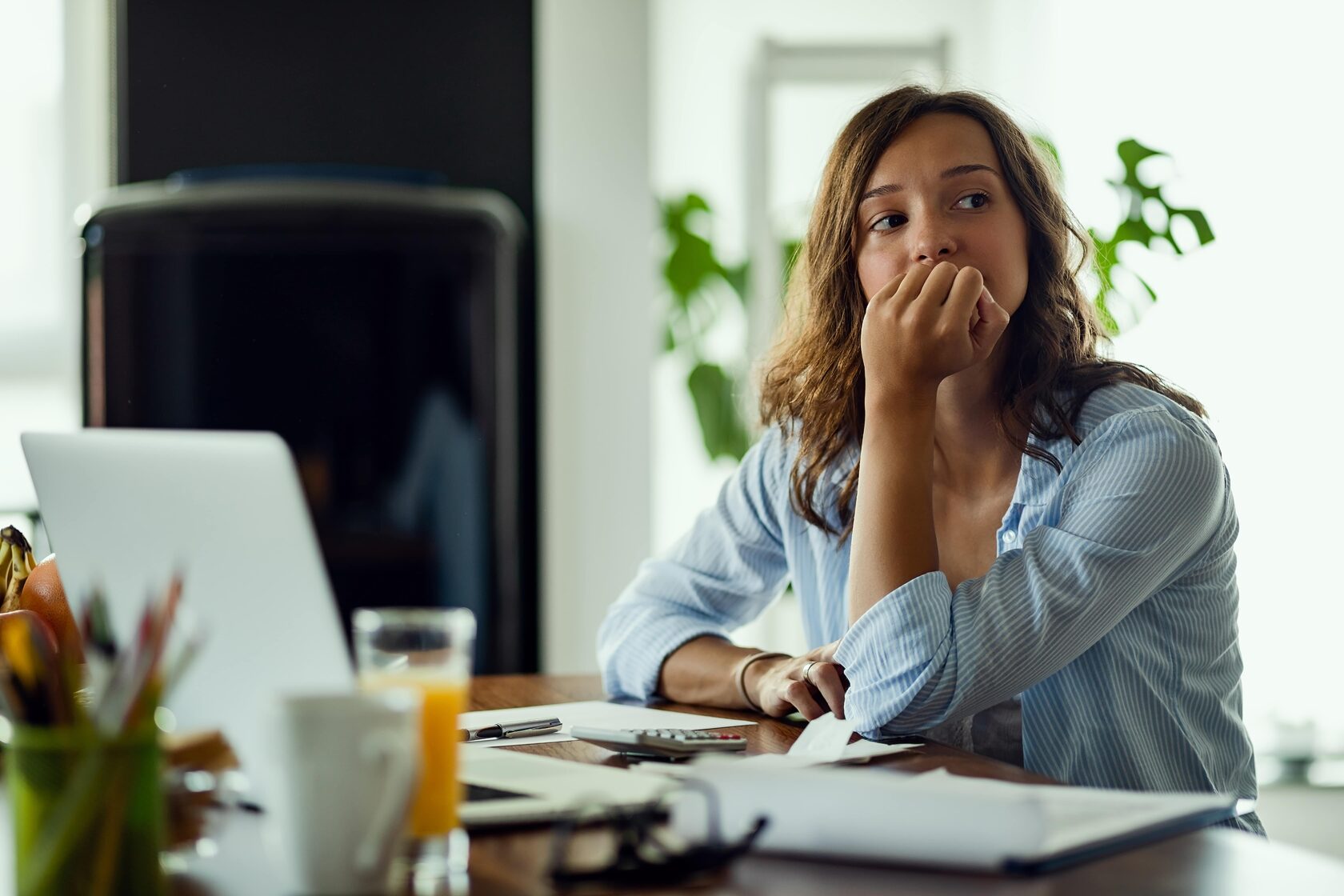 A young woman sitting at a desk with a laptop, papers, and a calculator, looking thoughtful and concerned. She appears to be deep in thought, possibly reflecting on job search challenges. Natural lighting and indoor plants add a relaxed yet contemplative