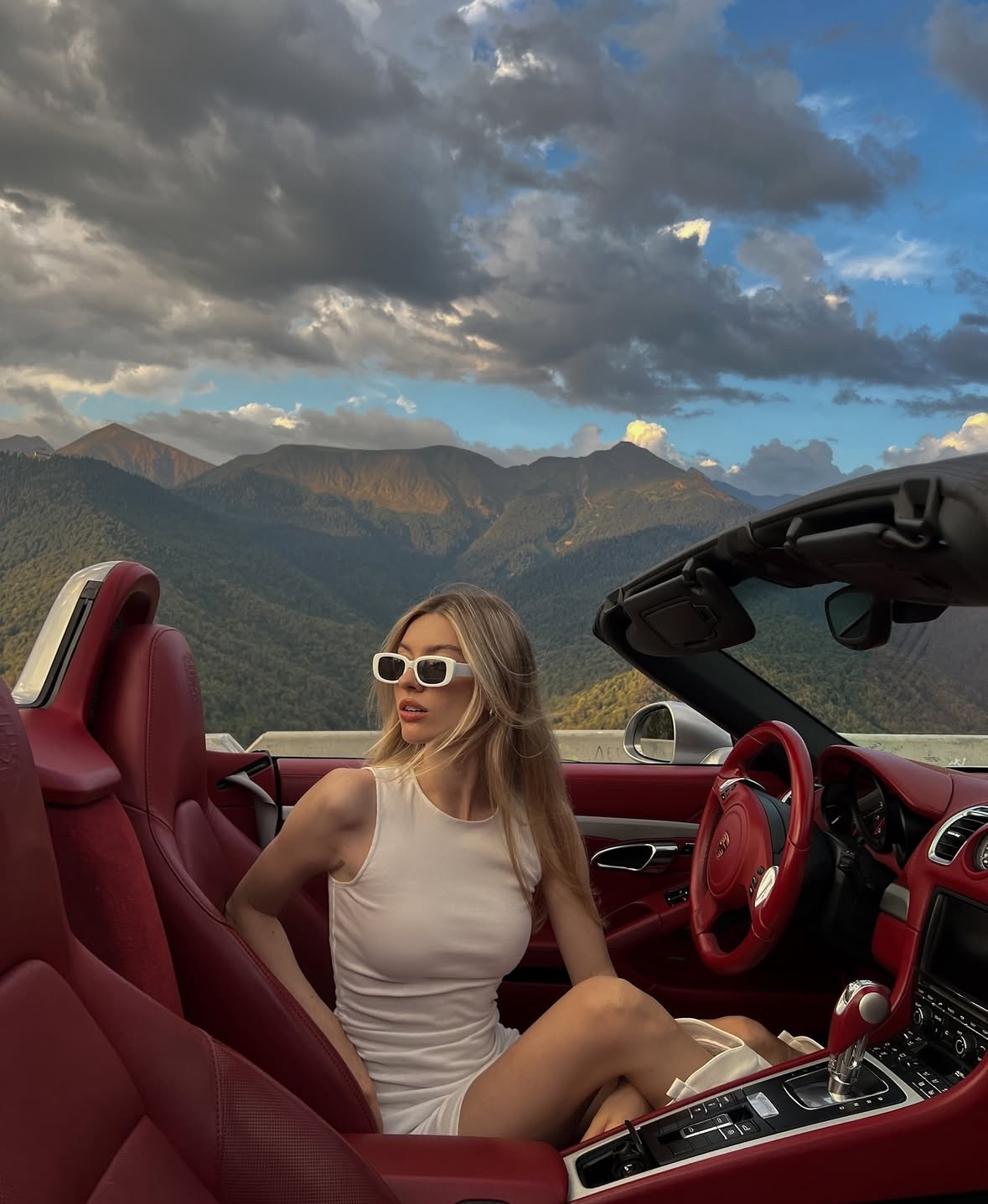 Девушка в открытом Porsche с красным салоном на фоне гор
