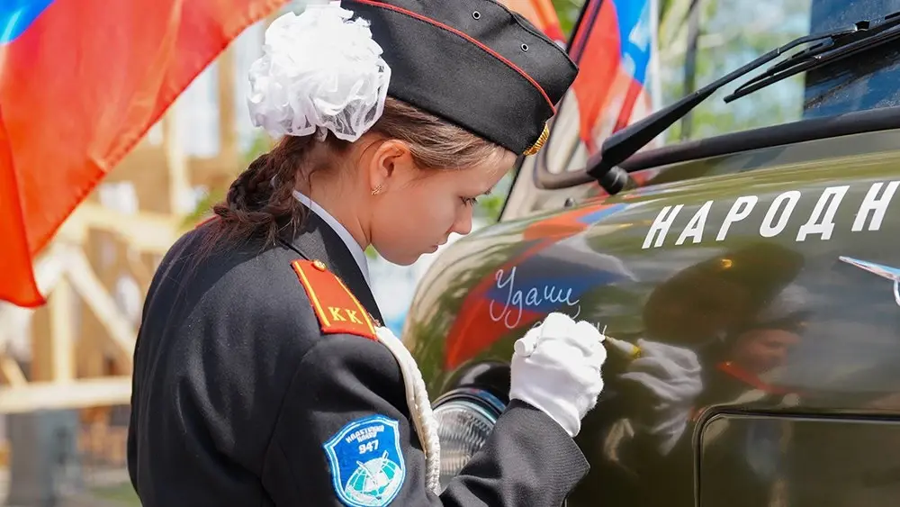 Девочка в форме пишет слова поддержки на автомобиле акции «Авто для фронта» в Национальном центре «Россия» ко Дню Победы.
