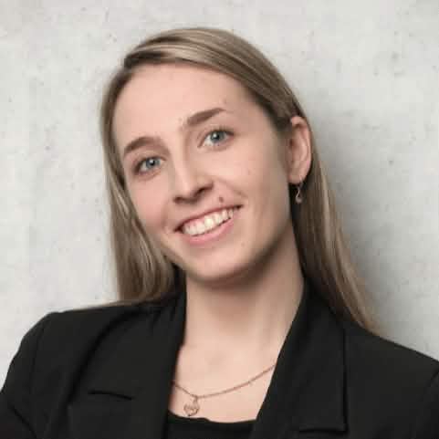 Портретная фотография молодой женщины со светлыми прямыми волосами, с лёгкой улыбкой и смотрящей прямо, в черном пиджаке и с золотистым кулоном на шее, на фоне однотонной серой стены.